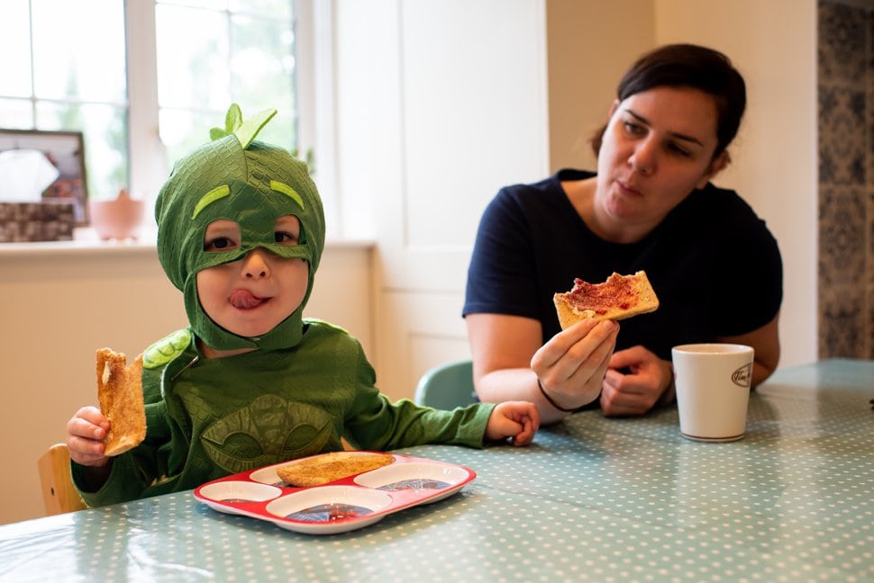 boy in PJ mask costume eats breakfast with tired mum