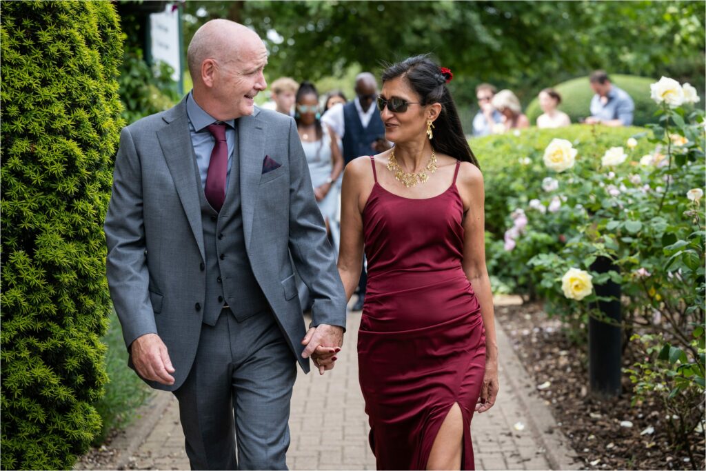 Bride wearing red dress, gold jewellery and sunglasses holds hands with groom in matching colour tie and enter St Albans Register Office