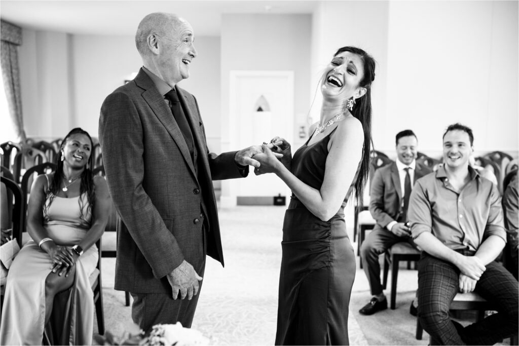 Bride laughs trying to get ring onto groom's finger during micro ceremony, guests laugh in background