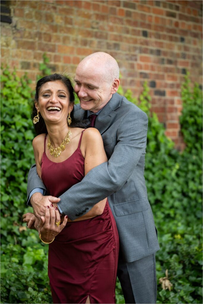 Groom hugs bride wearing red dress and gold jewellery as she laughs