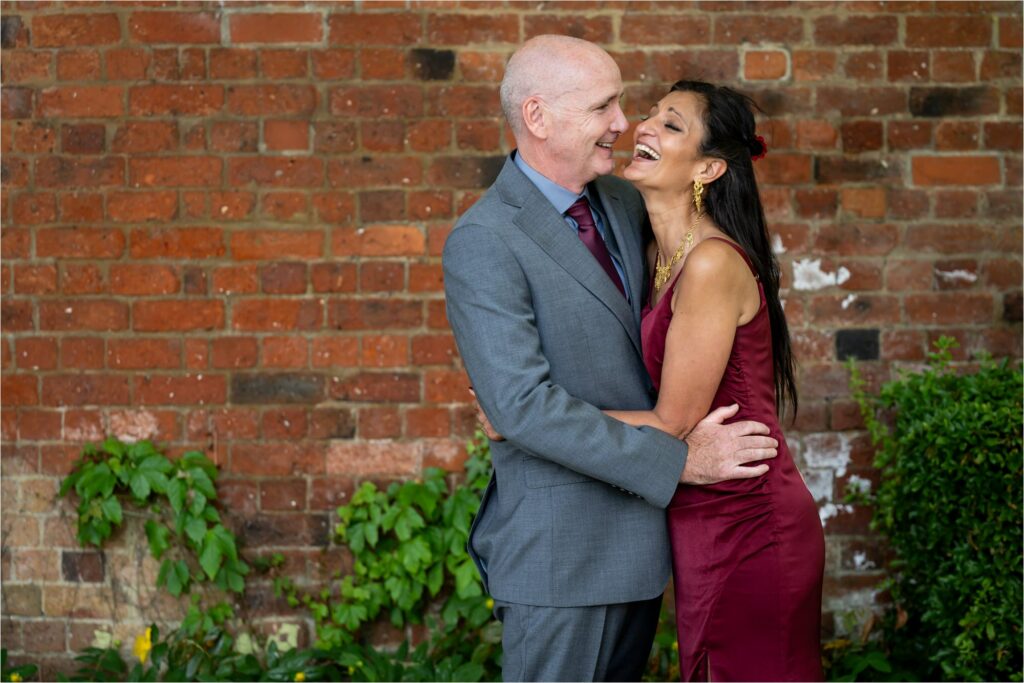 Groom makes bride in burgundy dress laugh against wall of St Albans Register Office