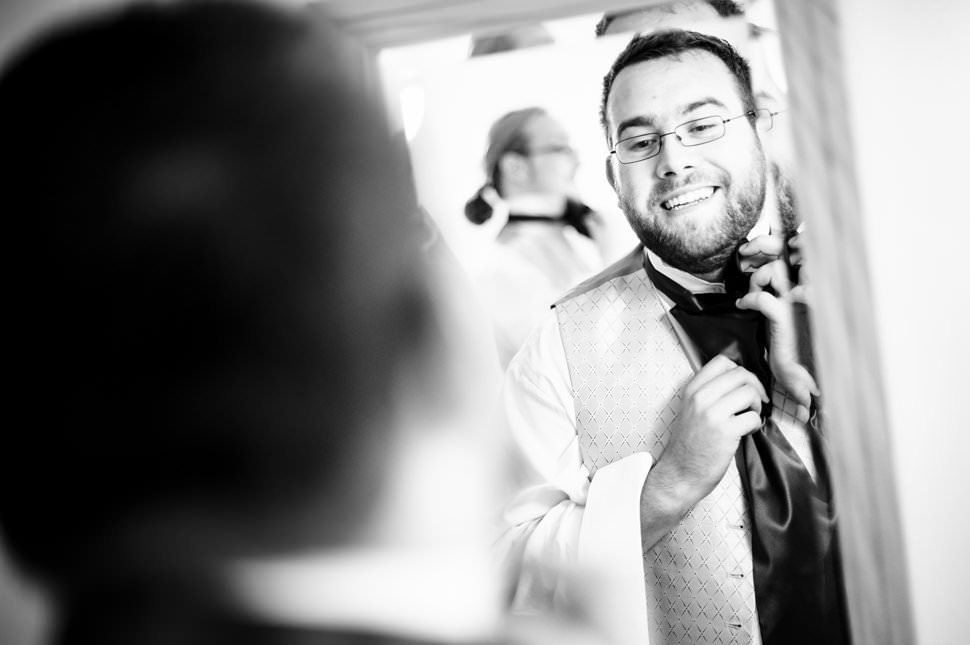 A black and white photo of a groom tying his cravat in a mirror before his wedding.