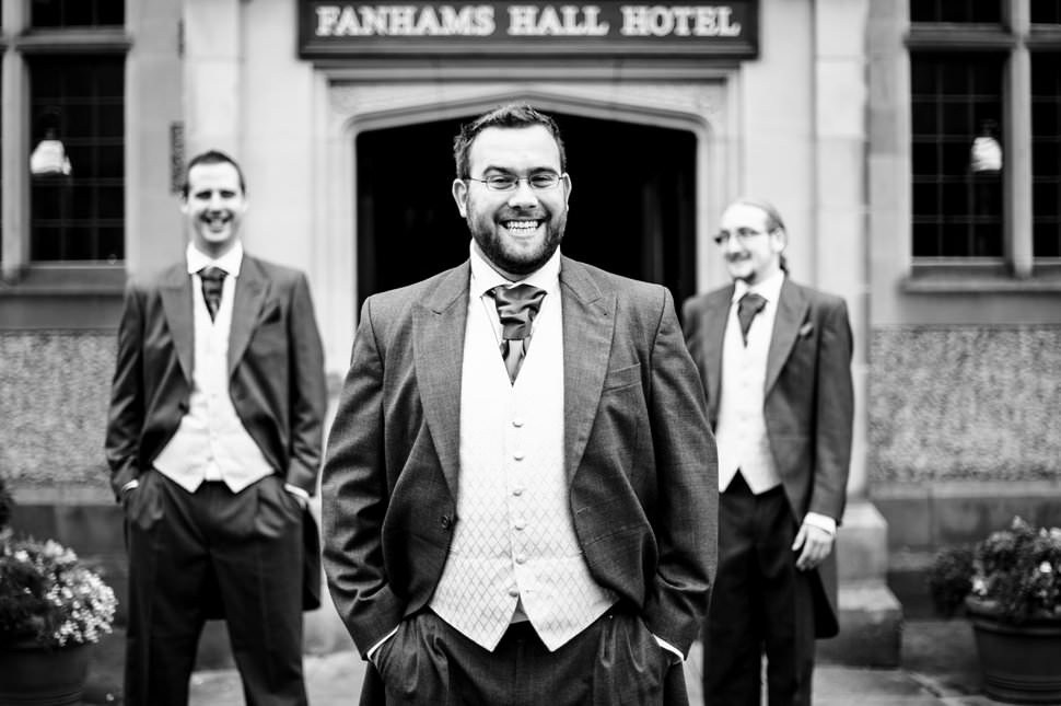 A laughing groom and his two groomsmen stand outside Fanhams Hall Hotel before their wedding.