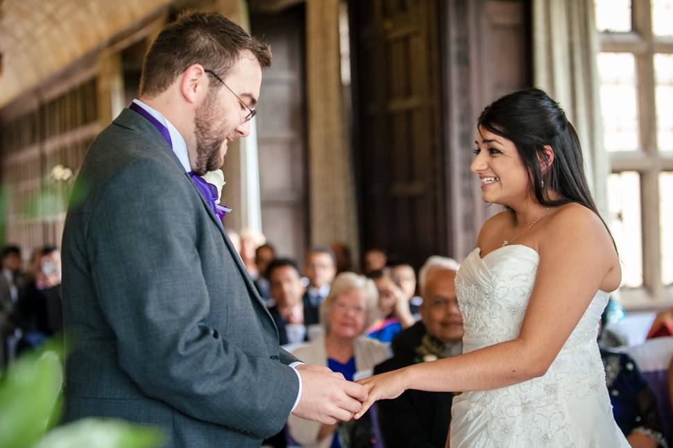 A bride laughs while the groom slips a ring onto her finger at this Fanhams Hall wedding in Hertfordshire.
