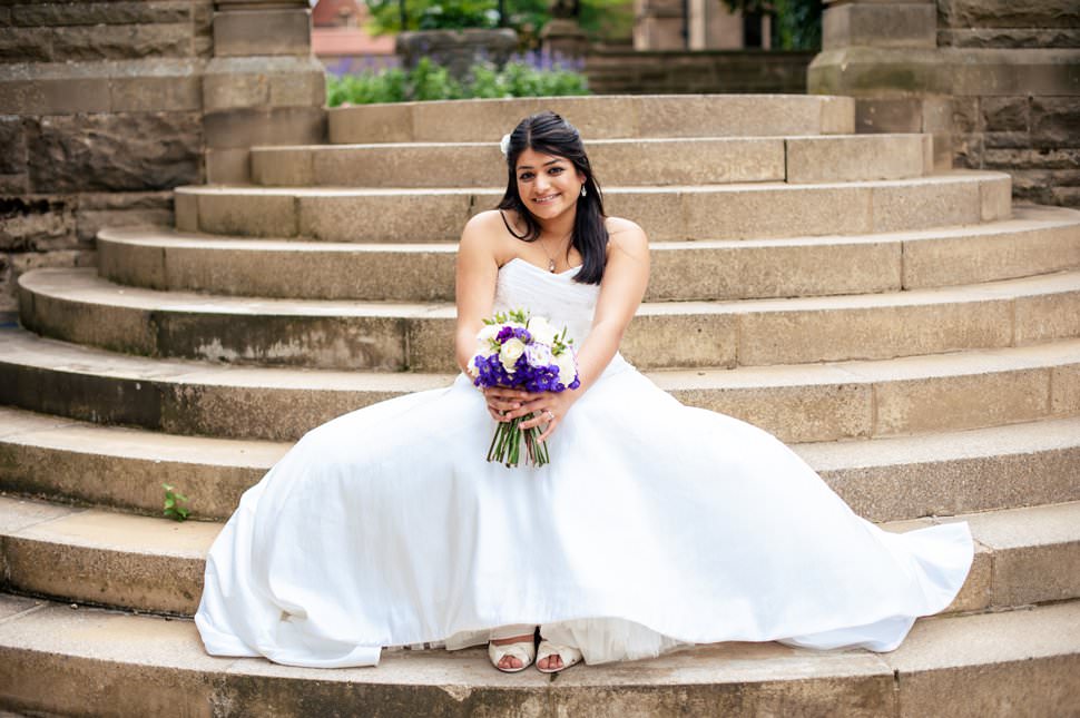 A bride sits on the beautiful steps at Fanham Hall holding a purple bouquet.