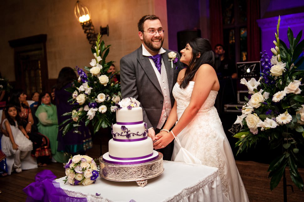 Bride and groom cut a white and purple wedding cake in front of large floral arrangements.