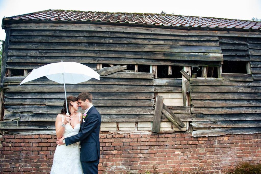 Bride and groom stand in front of run down barn under a white umbrella