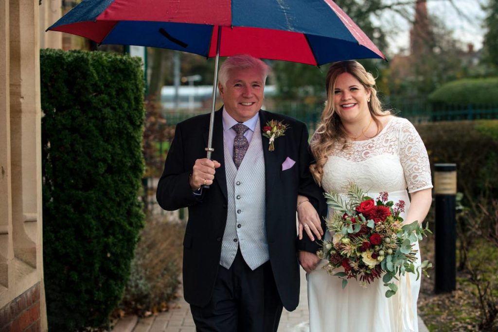 Bride enters St Albans Registry Office under colourful umbrella on arm of her father.