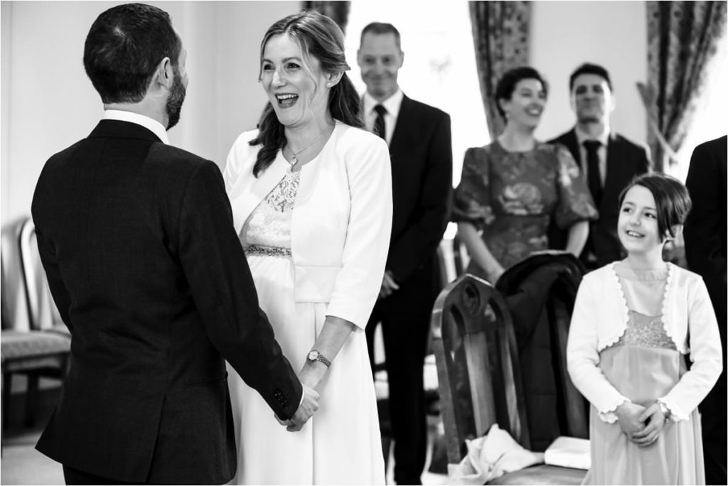 Bride laughs while holding groom's hands during St Albans Registry Office wedding ceremony.