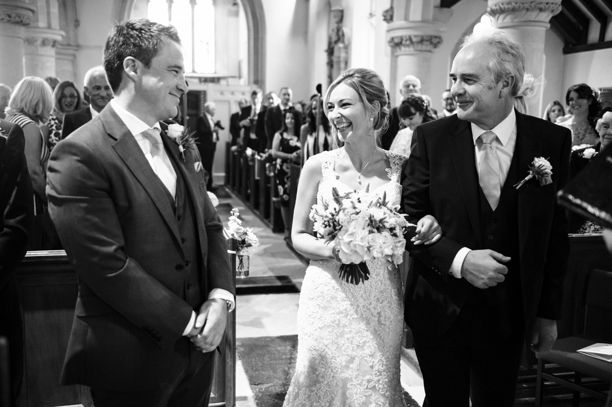 Bride and Dad smile at groom at top of church aisle