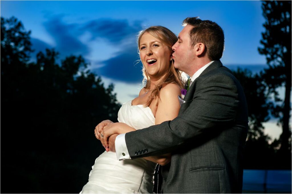 Groom kisses bride on cheek in front of evening blue sky