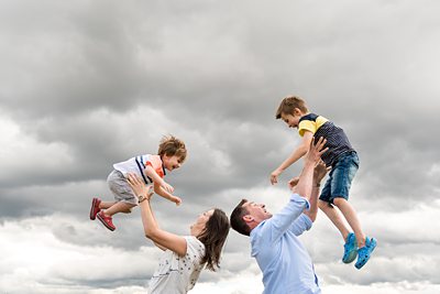 Happy family photo of parents throwing two sons in the air in front of cloudy sky in St Albans park