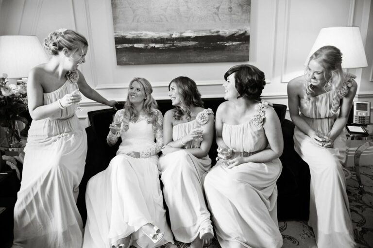 Bride laughs and drinks champagne with her four bridesmaids on the sofa