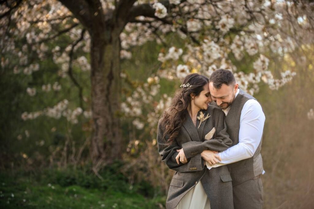 Bride wearing groom's suit jacket cuddles groom in front of cherry blossom tree at Essendon Country Club