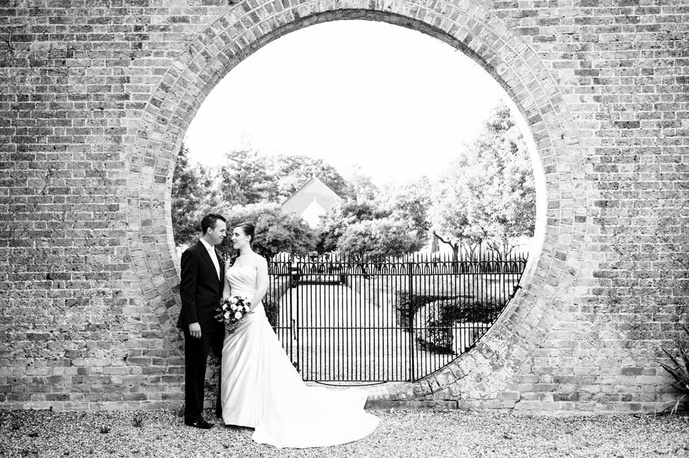 Couple stand in round hole in Hanbury Manor wall at Hertfordshire wedding
