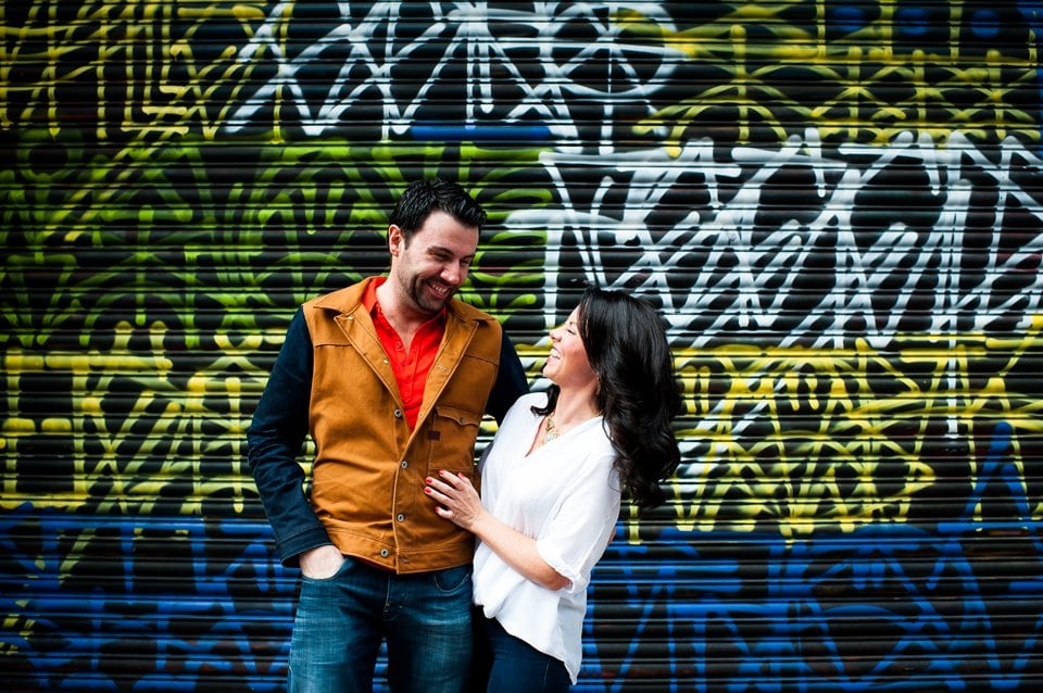 Couple stand in front of graffiti wall and laugh during engagement shoot
