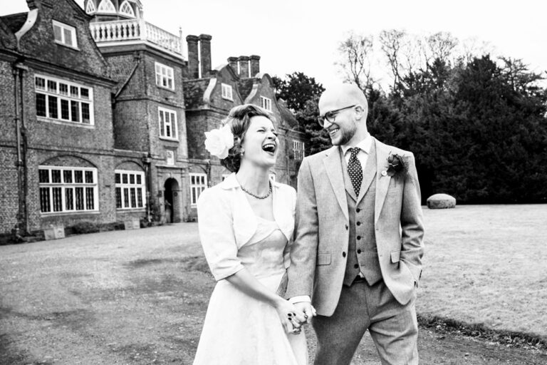 Laughing bride and groom walk in front of Rothamsted Manor in Hertfordshire