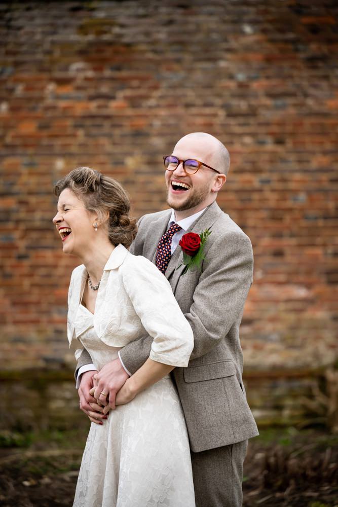 Laughing groom hugs laughing bride from behind in front of brick wall at Rothamsted Manor wedding