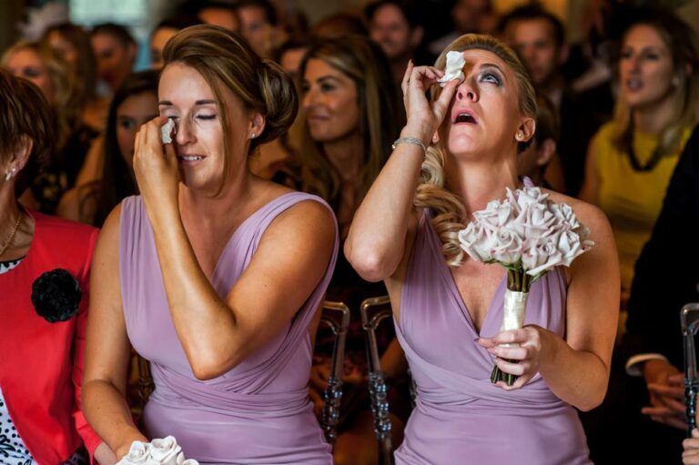 Two emotional bridesmaids wearing purple dresses wipe away tears during wedding ceremony