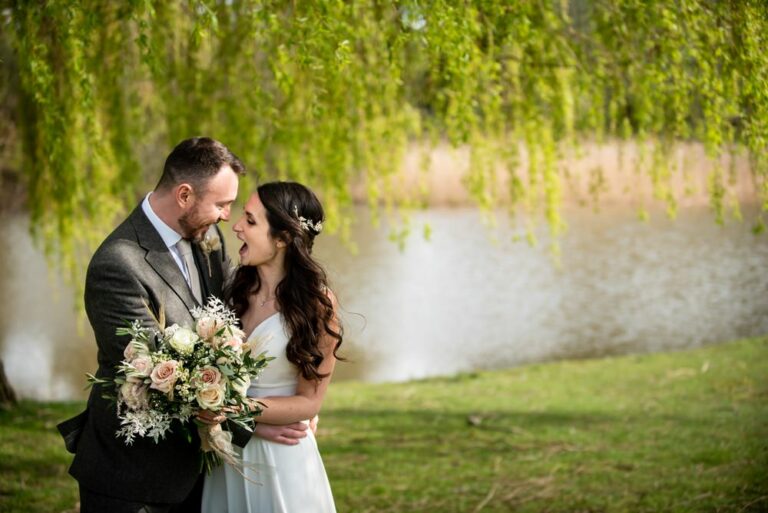 Happy couple snuggle in front of green weeping willow and lake at Essendon Country Club wedding