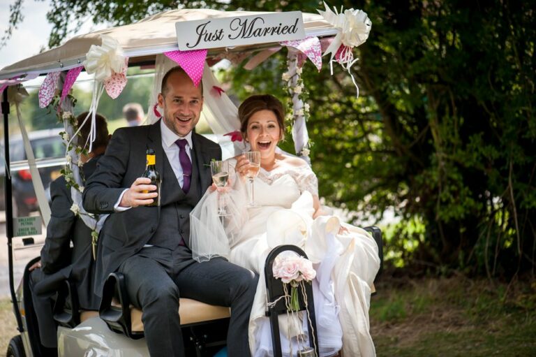 Laughing bride and groom drink champagne on just married golf buggy at Hertfordshire golf club wedding