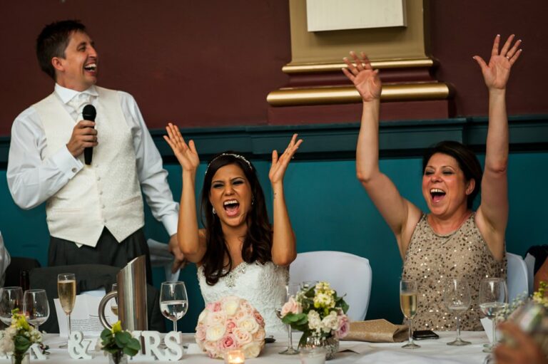 Bride and mother of bride cheer with hands in the air during groom's speech at Hertfordshire wedding