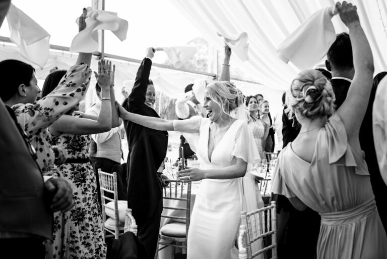 Bride walks into marquee wedding while guests wave napkins in the air at Hertfordshire wedding