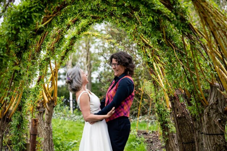 Two brides laugh during couple portraits under willow arch at South Farm wedding