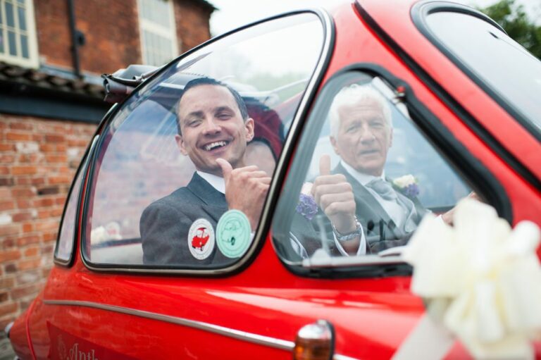 Groom gives thumbs up in red micro car with Dad on way to Hertfordshire wedding