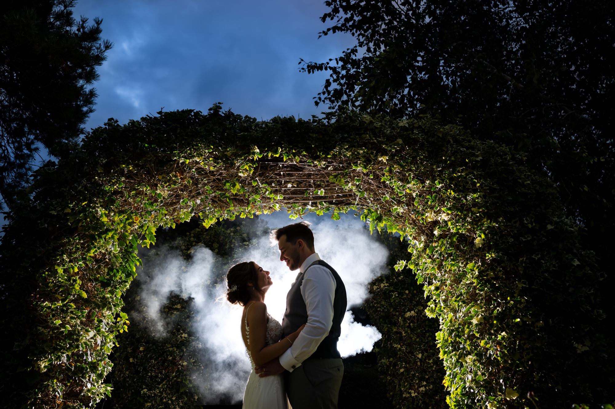 Nighttime shot of bride and groom in Tewinbury Farm tree arch backlit with smoke