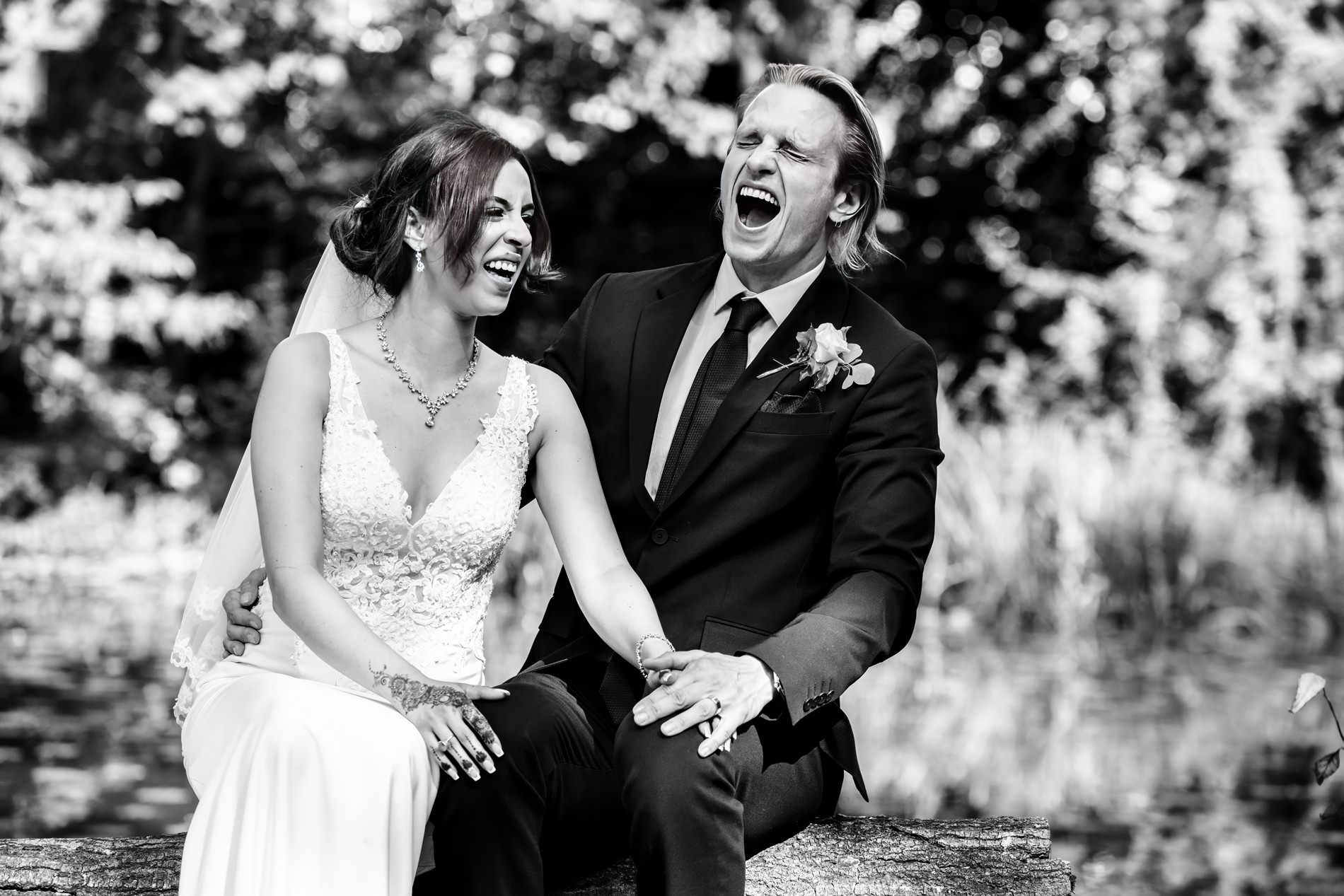 Bride and groom laugh while sitting on fallen tree trunk