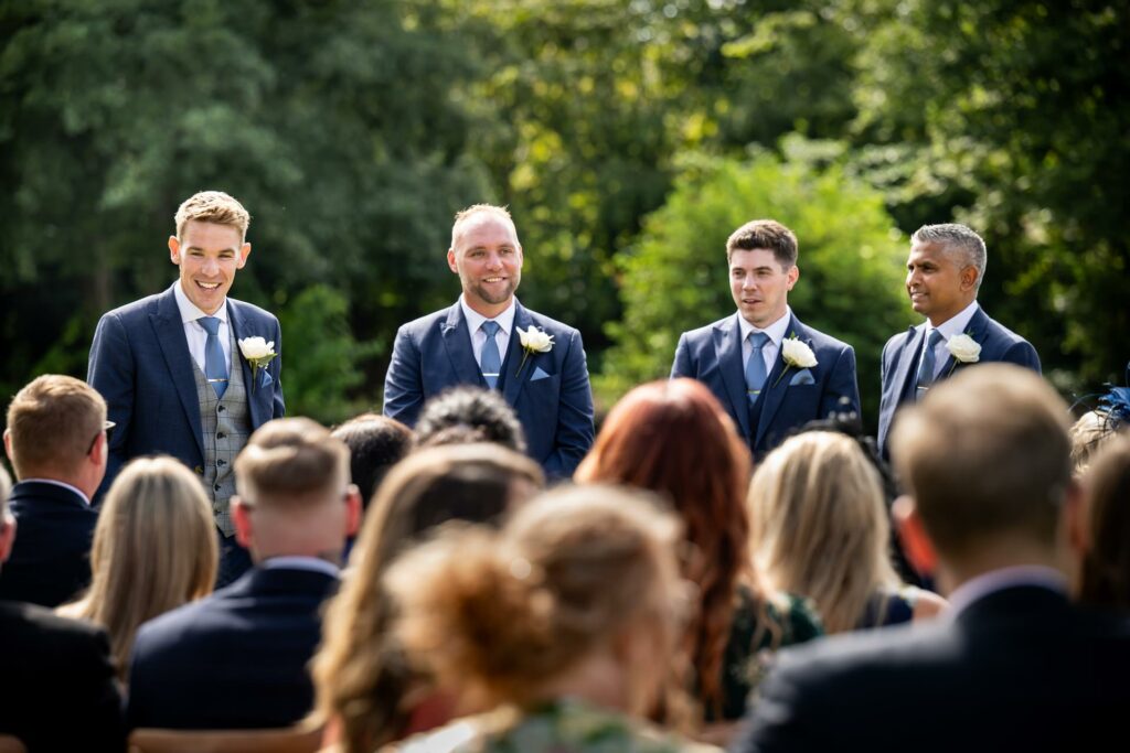 Groom and best men wait for bride behind sea of guests at St Michaels Manor outdoor wedding ceremony