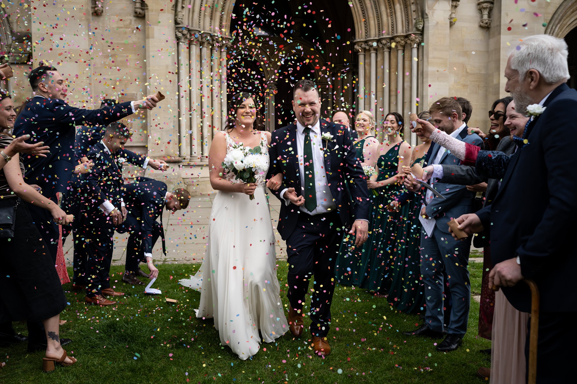 Bride and groom walk through coloured confetti cloud in front of St Albans Cathedral