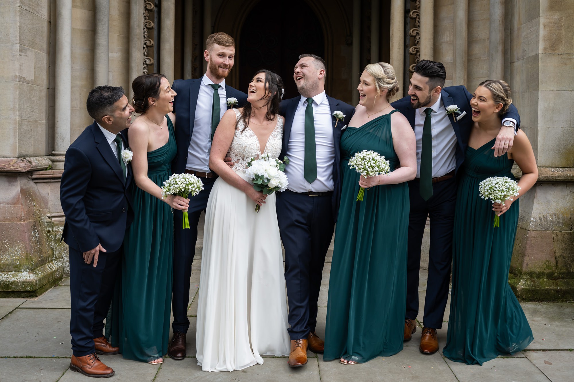 Bride, groom and wedding party in dark green pose for formal portraits and laugh in front of St Albans Cathedral