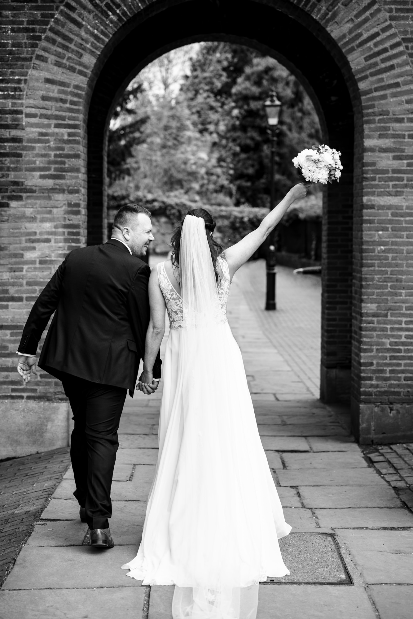 Bride throws flowers up walking through cathedral arch walking to pub reception after St Albans Cathedral wedding ceremony