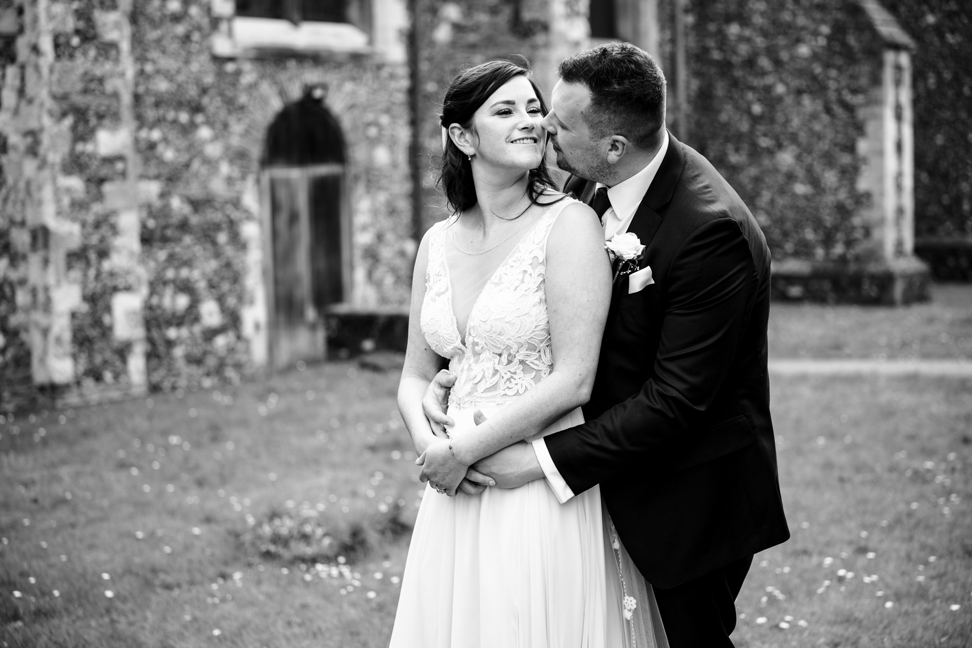 Groom hugs bride and kisses her cheek in grounds of St Albans cathedral, Hertfordshire
