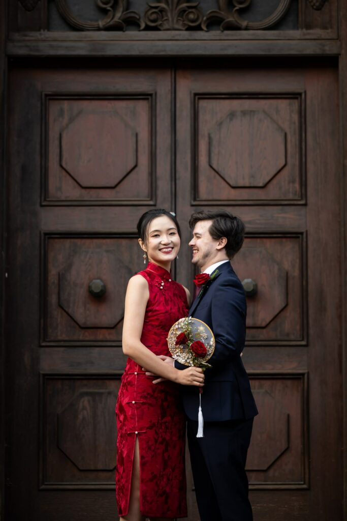 Bride in red dress and groom in suit stand by dark wooden door at Hertford Register Office