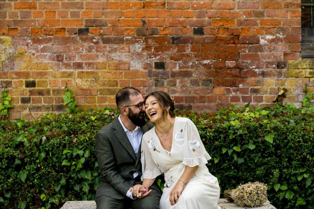 Bride and Groom sitting on bench laughing in St Albans Register Office gardens