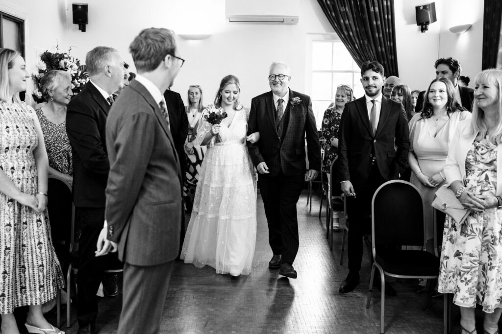 Bride is walked down aisle by father during Hertford Register office intimate wedding in black and white photo