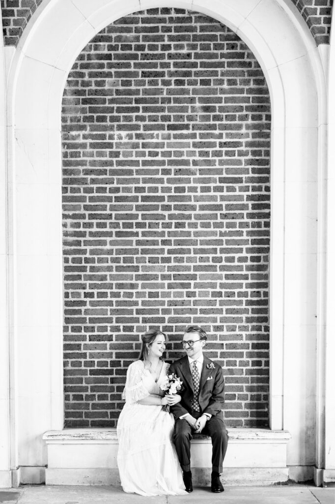 Bride and groom sit on bench in white arches at Hertford Register office