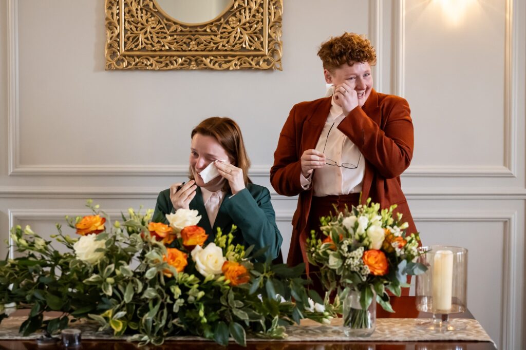 Two brides cry on signing the marriage register at the St Albans Register Office behind orange and white DIY flowers and bouquets