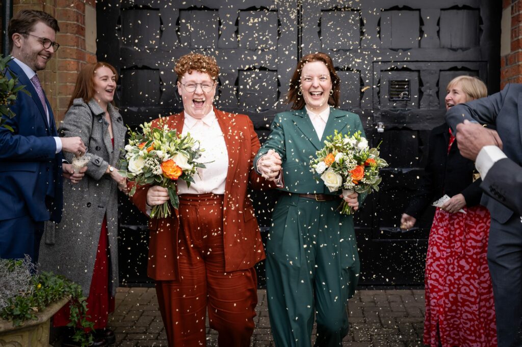 Two brides laugh in a cloud of white confetti on leaving the St Albans Register Office after their wedding ceremony