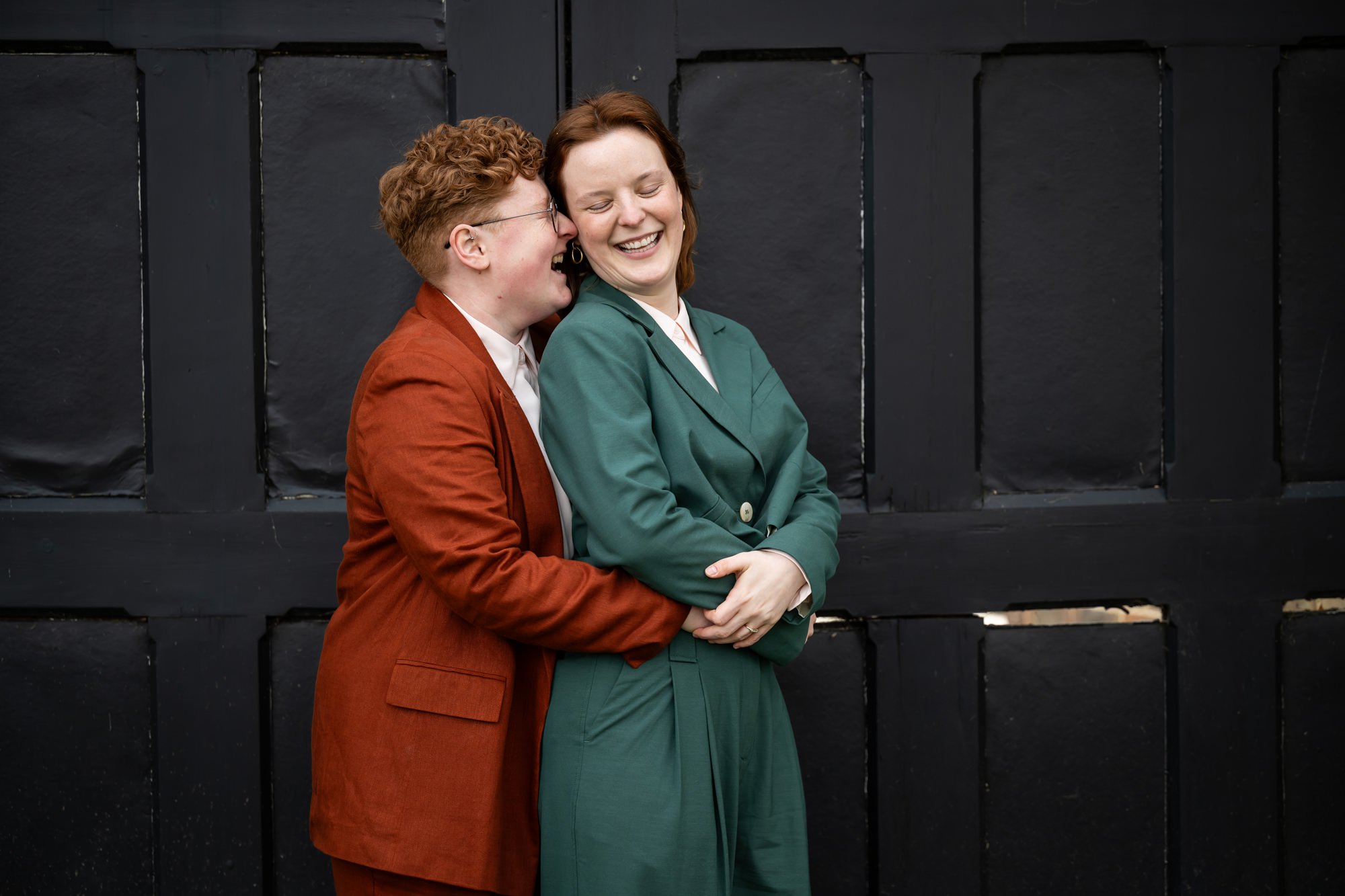 Two brides laugh in front of the St Albans Register Office black doors as one whispers in the other's ear