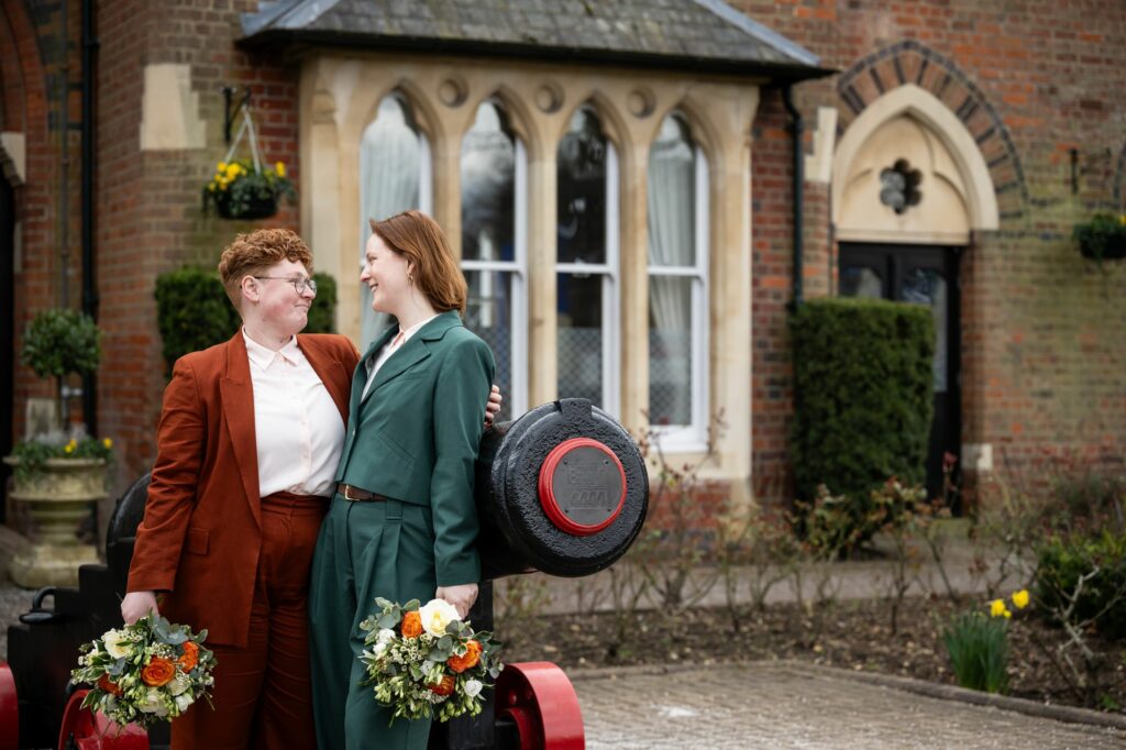 Two brides wearing suits and holding flowers lean on the St Albans Registry Office canon and gaze adoringly at each other with the building in the background at a winter wedding