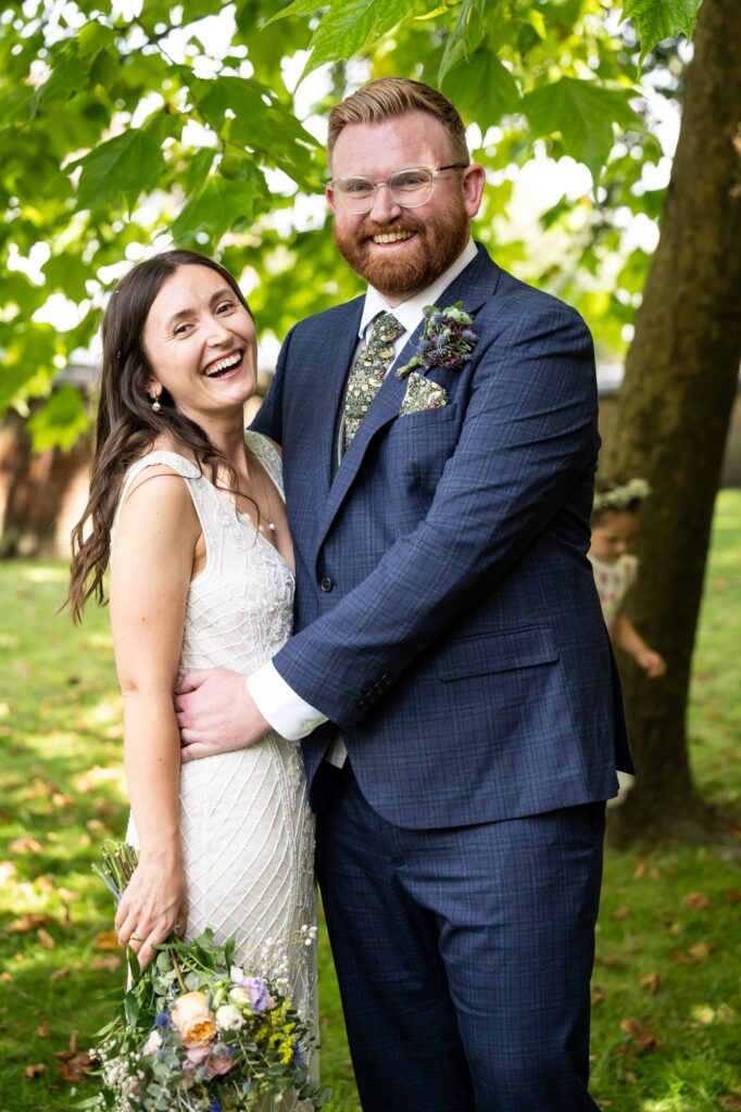 Bride and groom stand under trees during summery couple portraits at St Albans Registry Office wedding