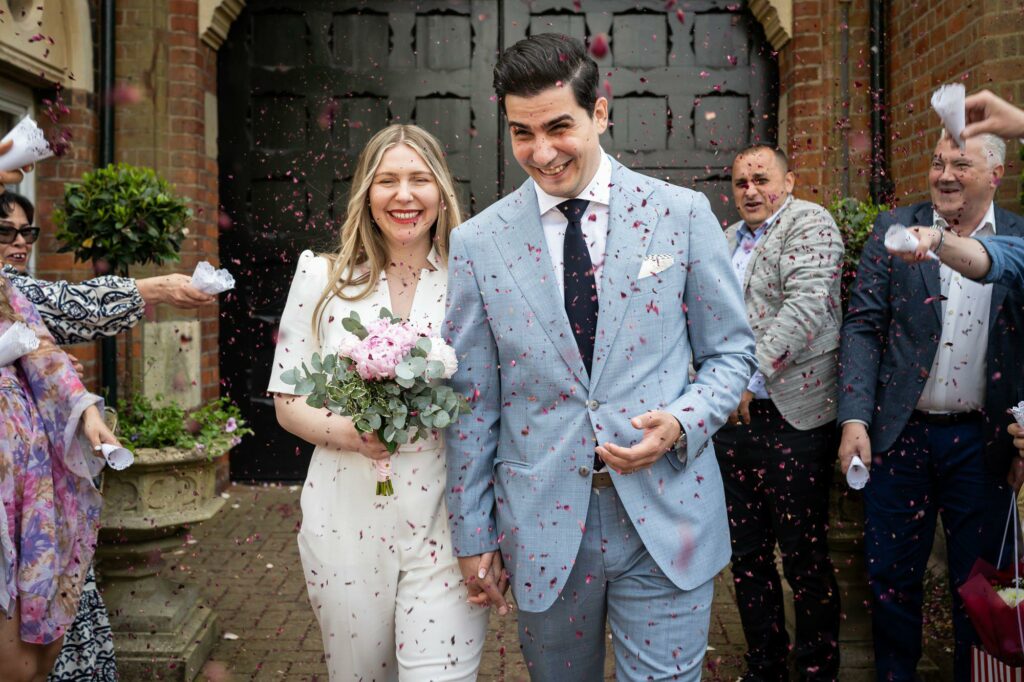 Bride in white jumpsuit and groom in grey suit exit St Albans Registry Office in cloud of pink confetti after micro wedding ceremony