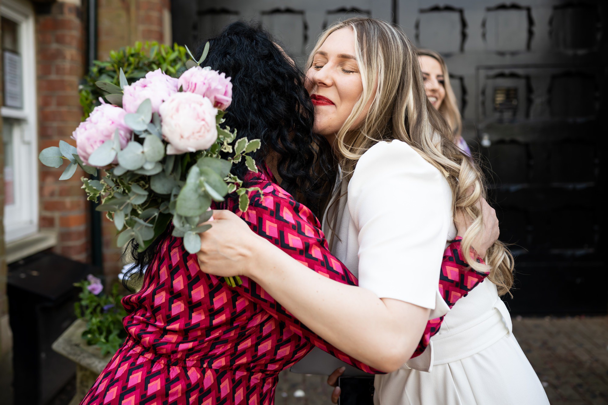 Bride embraces friend outside St Albans Register Office holding wedding bouquet of pink peonies