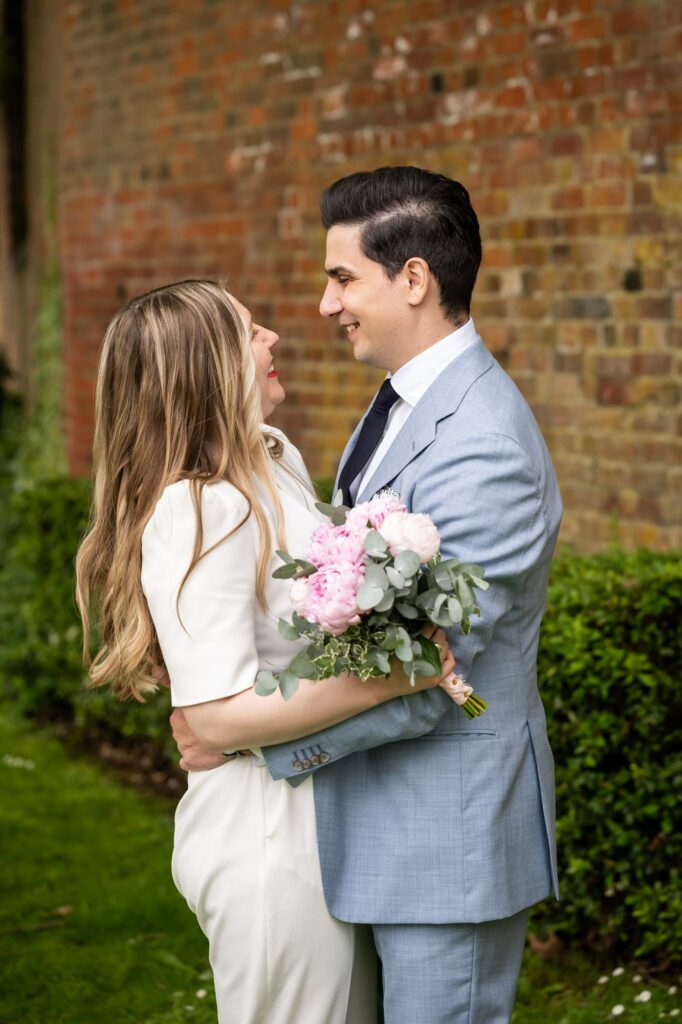 Bride in white jumpsuit with pink peonies and groom in grey suit embrace outside St Albans Registry Office