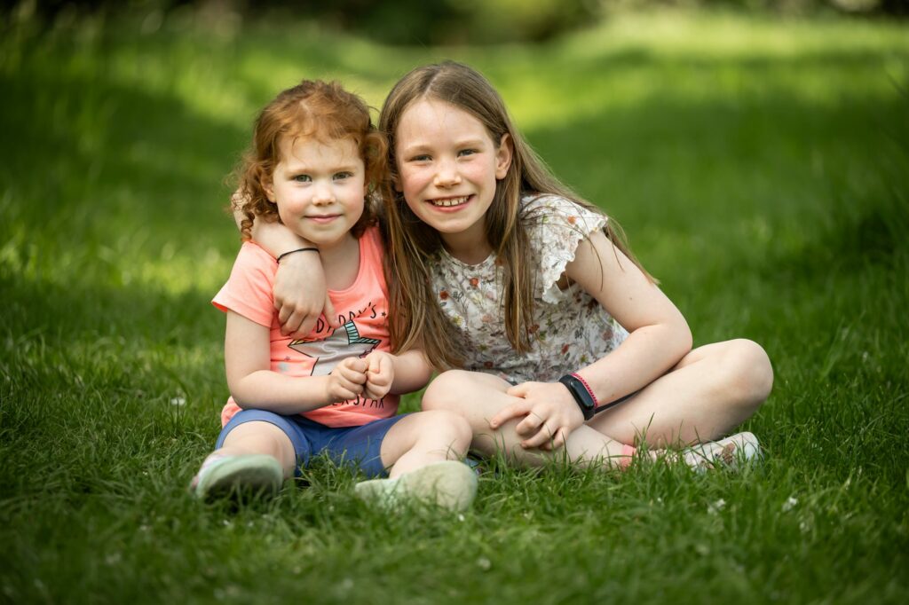 Two girls sit in grass smiling at camera and hugging during family photoshoot in St Albans
