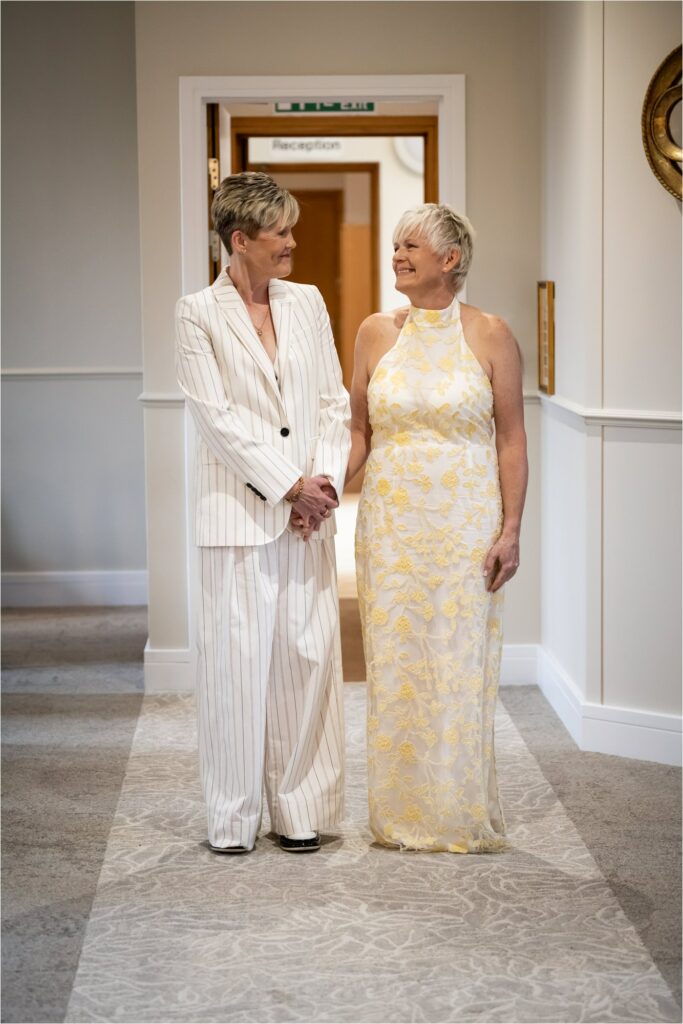 Two brides enter room wearing white pinstripe suit and white patterned dress for their civil partnership ceremony.